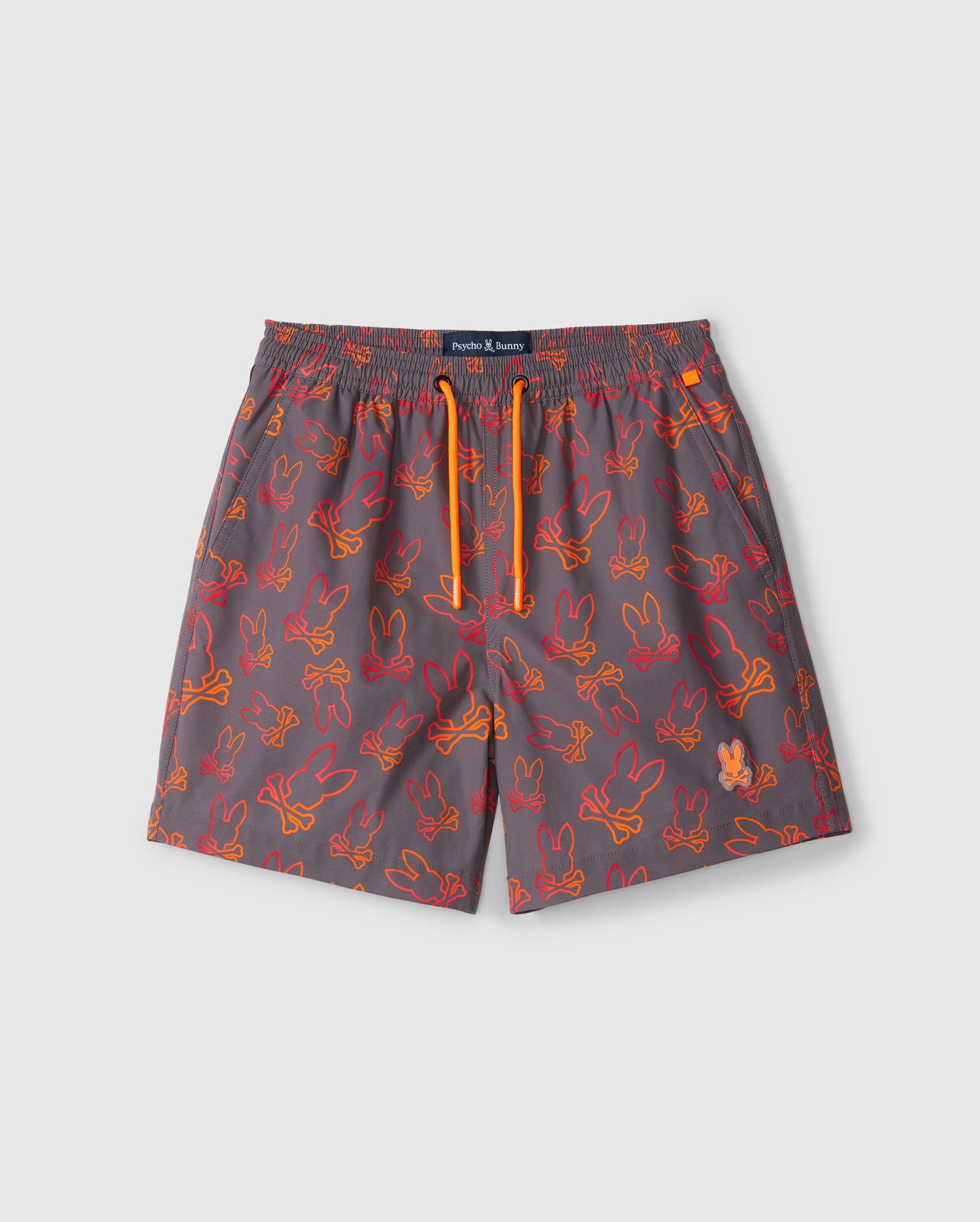 Gray kid's swim trunks featuring an all-over playful bunny pattern in orange and red outlines. These shorts have a quick-dry fabric, an elastic waistband with a bright orange drawstring, and a navy label on the waistband. The trunks are the KIDS SHELDON ALL OVER PRINT SWIM TRUNK - B0W588C200 by Psycho Bunny.