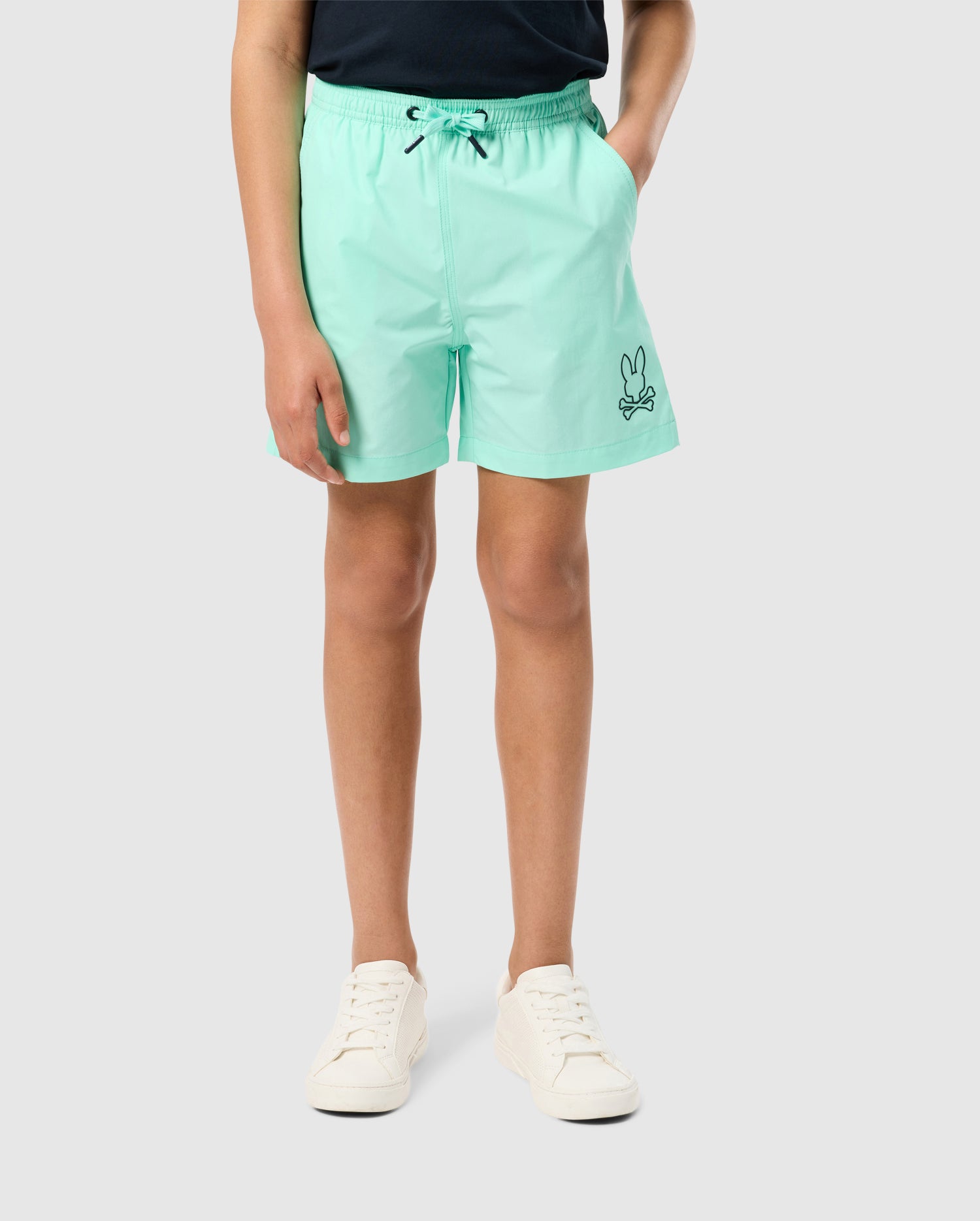 A person is standing against a plain background, wearing mint green KIDS PARKER HYDROCHROMIC SWIM TRUNK - B0W646C200 from Psycho Bunny with an embroidered bunny logo on the left leg, a black drawstring, and a dark top. They also wear white sneakers. Their hands are in the pockets of the trunks.