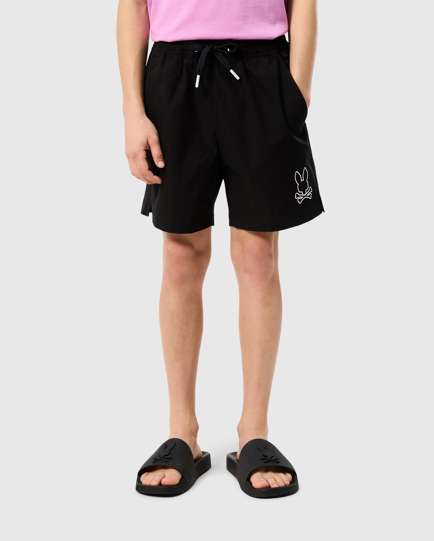 Person wearing a pink shirt, black Psycho Bunny KIDS PARKER HYDROCHROMIC SWIM TRUNK - B0W646C200 with a small bunny graphic on the left leg, and black slide sandals. The quick-dry shorts have pockets, and the individual is standing against a plain white background.