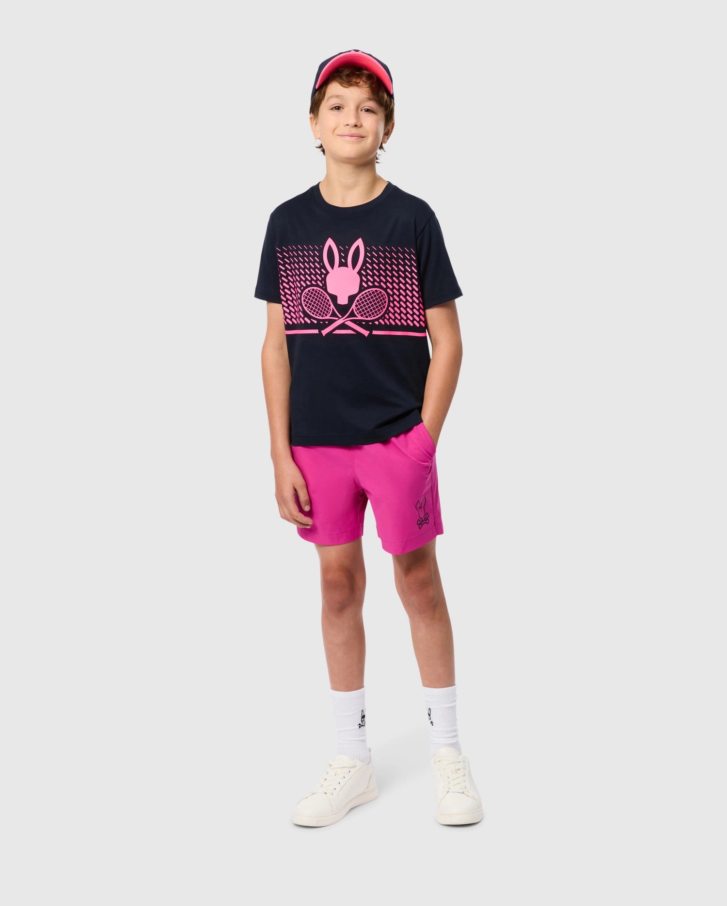 A young person stands confidently with hands in pockets, donning a navy T-shirt featuring a pink bunny pattern and crossed tennis rackets graphic. They sport the bright pink, quick-drying **KIDS PARKER HYDROCHROMIC SWIM TRUNK - B0W646C200** from **Psycho Bunny**, paired with white socks, white sneakers, and a pink visor against a plain light grey background.