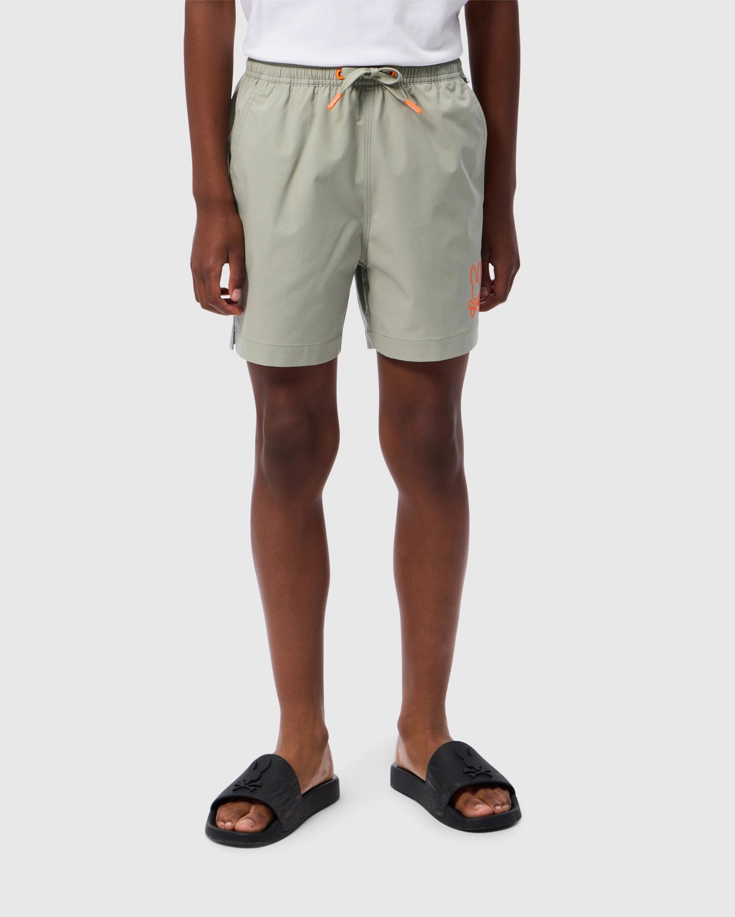 A person is wearing light green Psycho Bunny KIDS PARKER HYDROCHROMIC SWIM TRUNK - B0W646C200 with an orange logo on the left leg, and black slide sandals. The background is plain white. The person’s upper body is not visible.