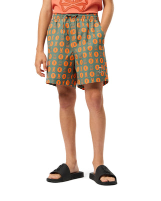 A person in an orange shirt and Kids Arnold Print Swim Trunk by Psycho Bunny, featuring a green design with an orange pattern, stands against a white background. The quick-dry material and stretch mesh lining of the trunks are complemented by black slides.