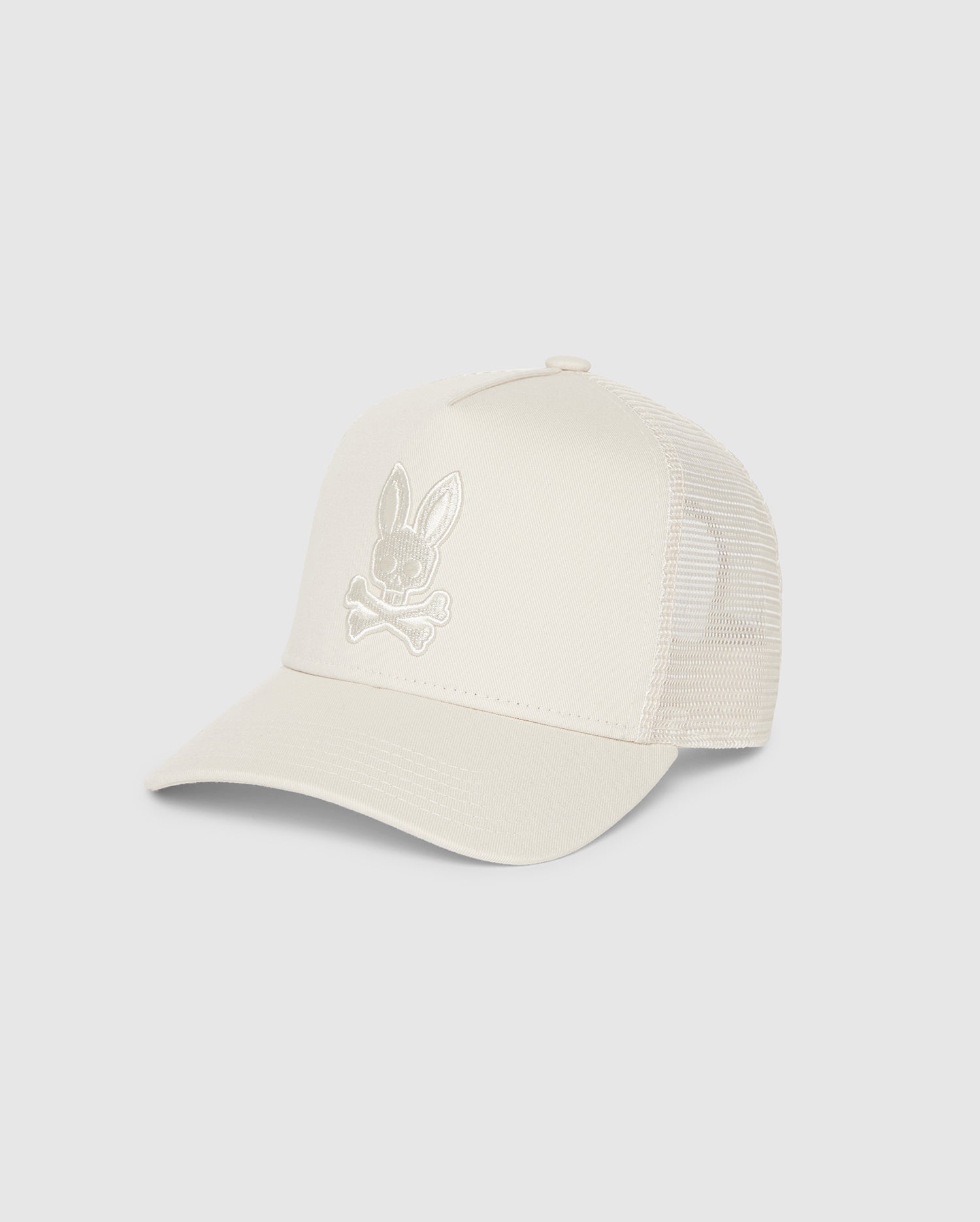 A beige men's trucker cap with a curved brim and mesh back. The front features a tonally embroidered Bunny design above two crossed bones, all in a similar beige color, providing a subtle yet distinctive look. It also includes snapback fastening for an adjustable fit. The Psycho Bunny MENS JAY TRUCKER CAP - B6A123B2HT is perfect for adding style to any casual outfit.