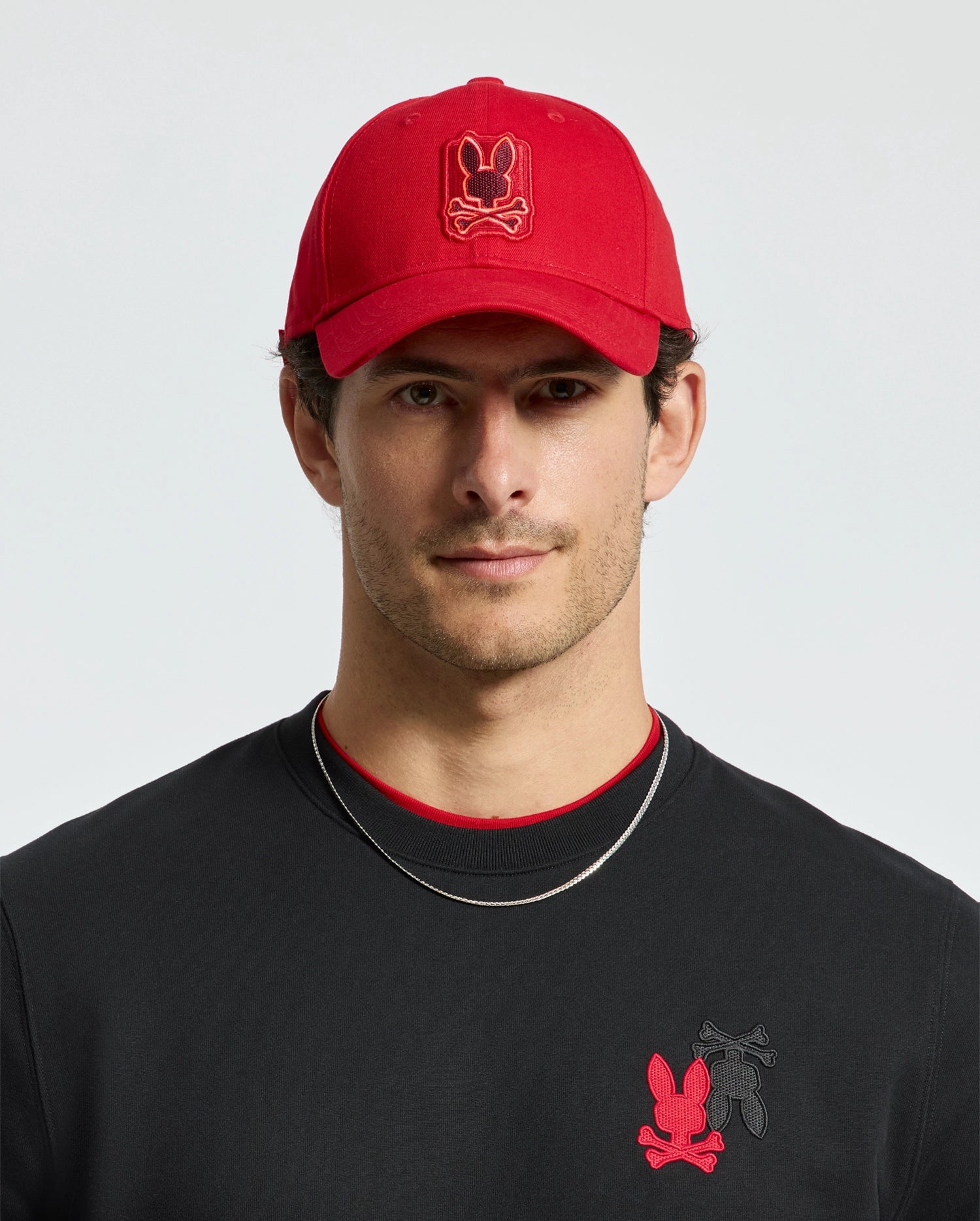 A man with a neutral expression wears the stylish Psycho Bunny MENS PIERCE BASEBALL CAP - B6A183E200, adorned with a cartoon bear design. His black shirt also features the same red and black bear theme, perfectly matching his short dark hair and thin silver necklace, crafting an outfit that embodies cotton comfort.