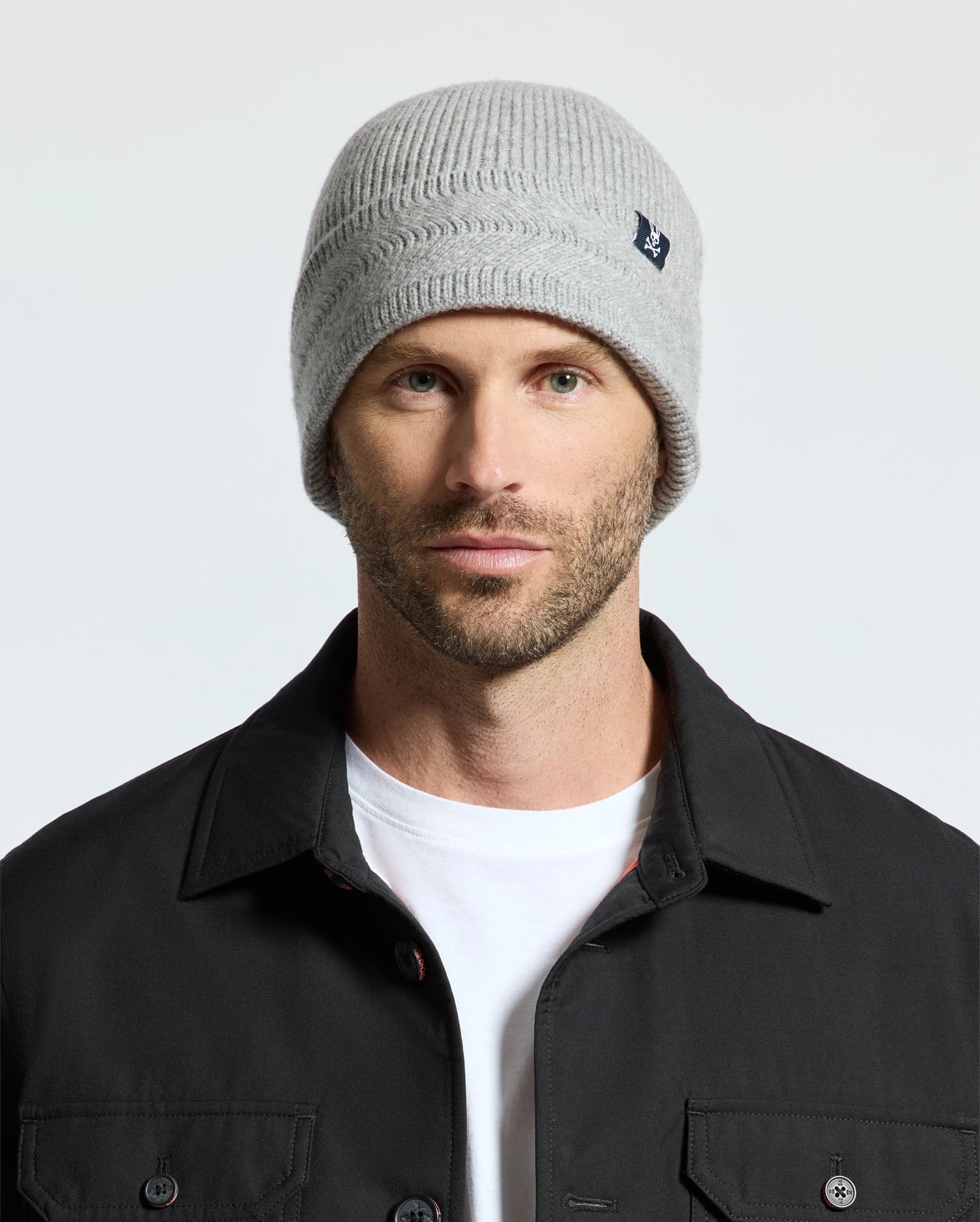 A man wearing a Psycho Bunny MENS RACKED BEANIE (B6A188E200) and a black jacket over a white t-shirt stands against a plain background.