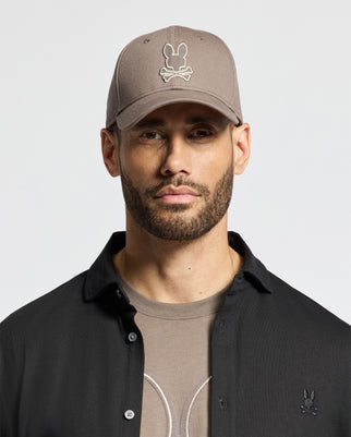 A man with a beard wears the Psycho Bunny Men's Alexander Baseball Cap in taupe, showcasing the brand's iconic bunny logo above crossed bones. He is dressed in a black button-up shirt layered over a taupe T-shirt that has a subtle design. The premium quality of the hat stands out against the plain white background.