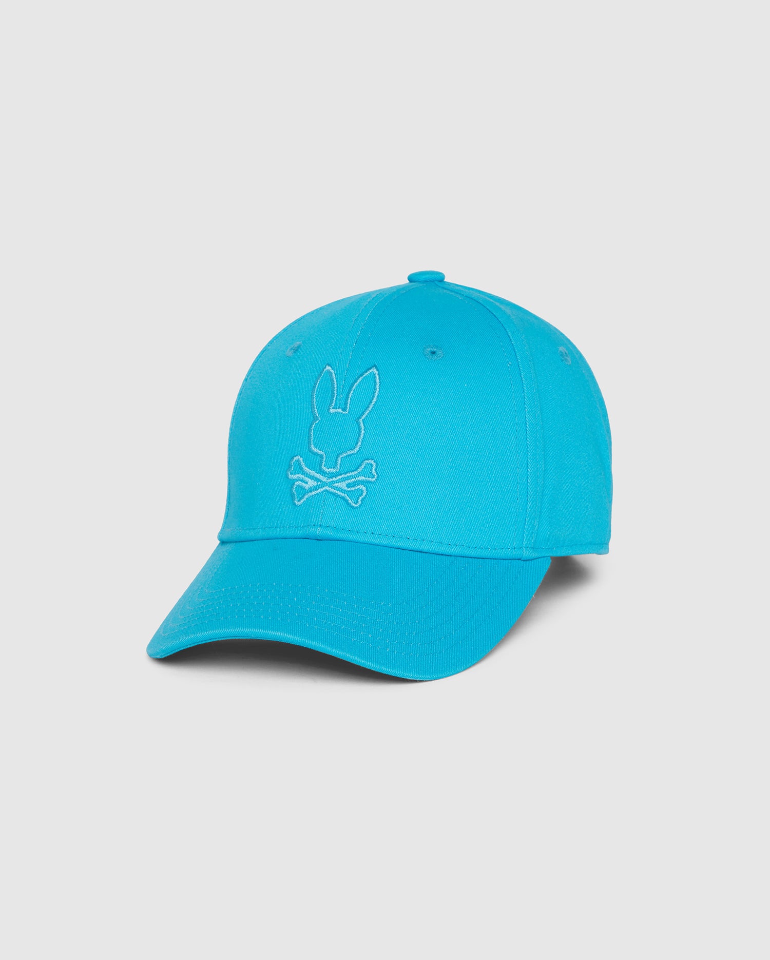 A bright blue Psycho Bunny MENS DANBY BASEBALL CAP - B6A360B2HT with a curved brim and an embroidered design on the front. The design features a Bunny head above a pair of crossed bones. The cap has a button on top and is displayed against a plain white background.