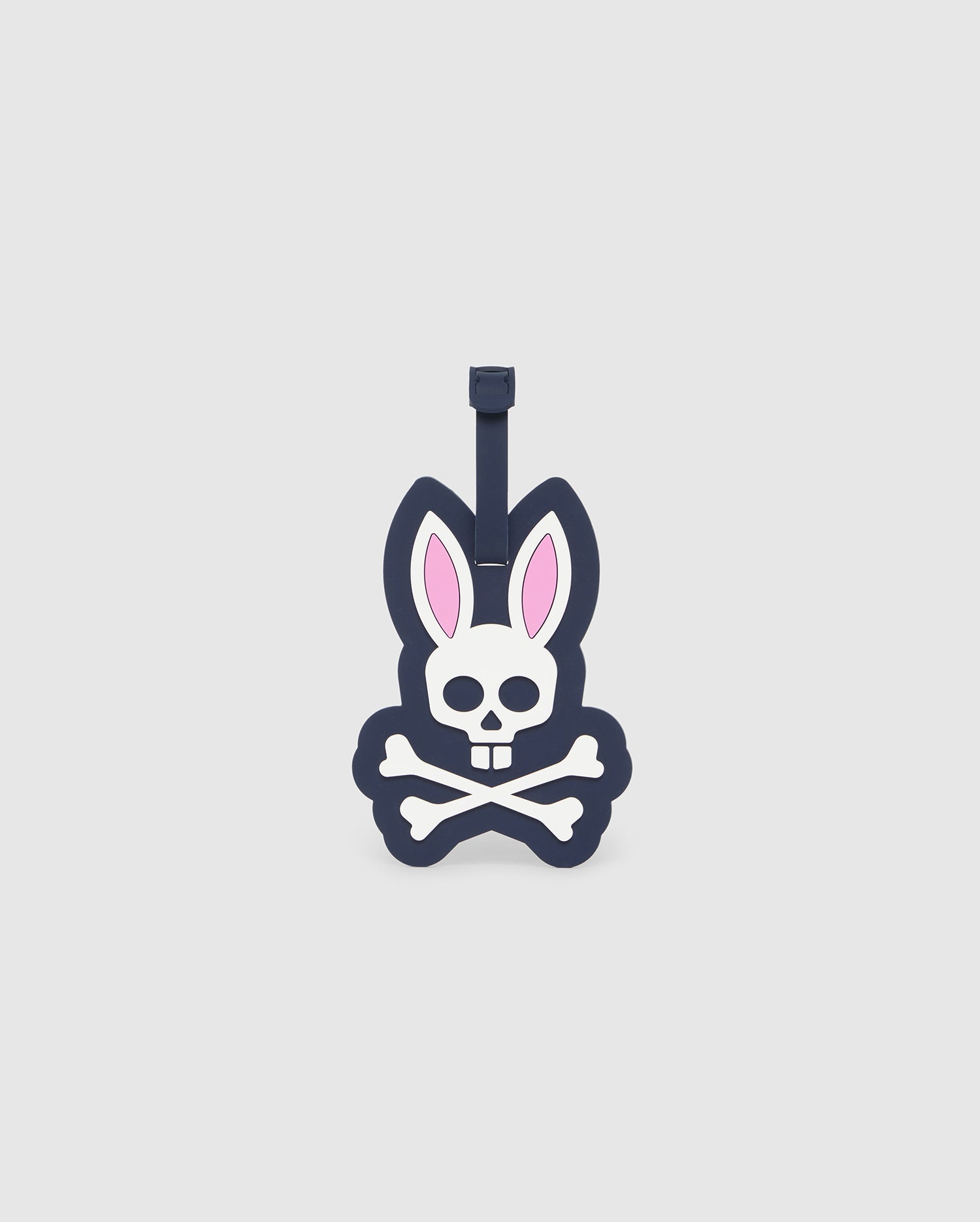 The Psycho Bunny MENS LUGGAGE TAG - B6A387B200 is a detachable silicone luggage tag featuring a cartoon design of a skull with rabbit ears and crossbones underneath. The skull has a simple outline with a concealed bunny nose, with the ears in white and pink. The tag is primarily dark blue and white, complete with a personal information slot for added convenience.