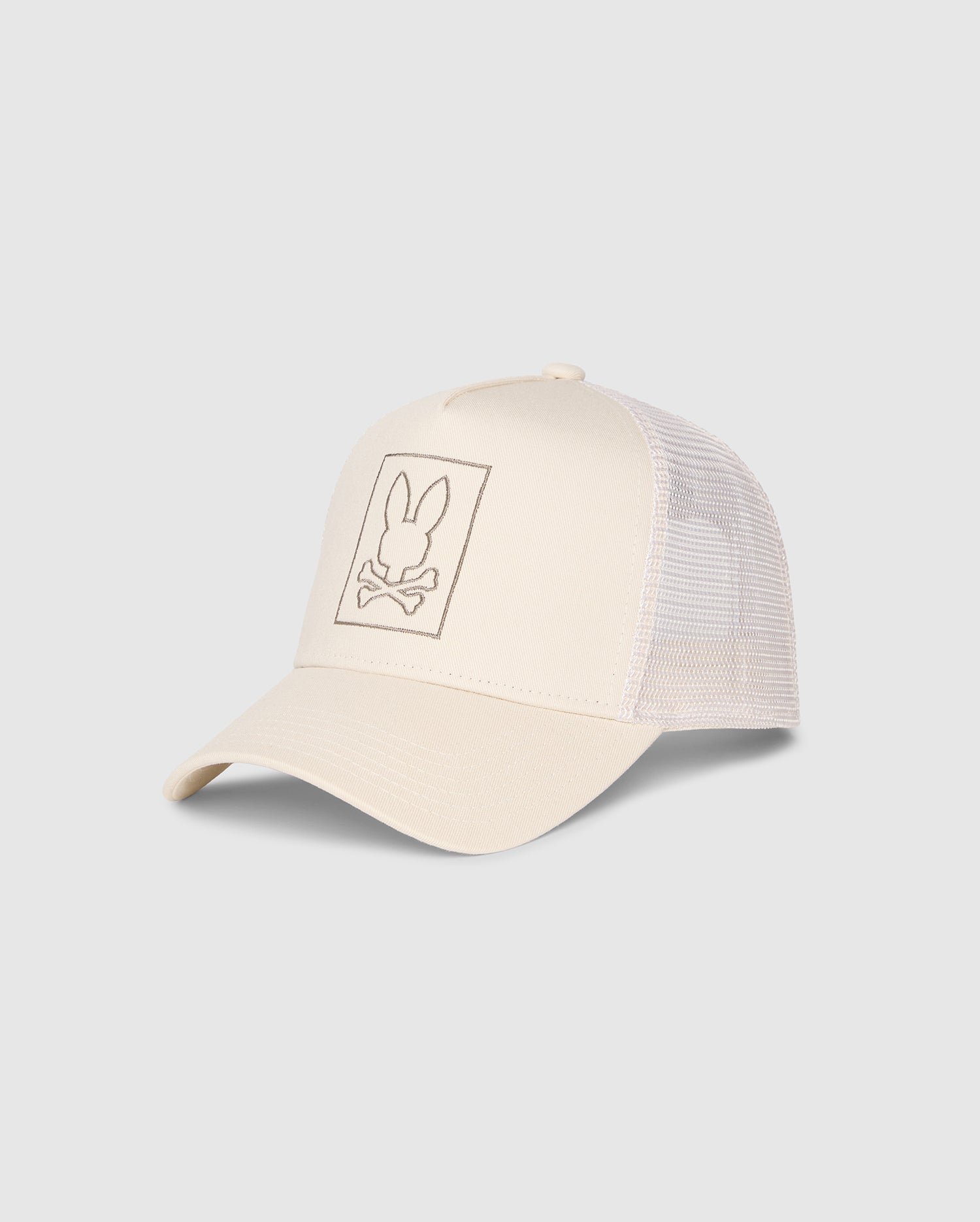 The Psycho Bunny MENS LIVINGSTON TRUCKER CAP - B6A401B200, in beige, features an adjustable snapback and mesh back. The solid front showcases a white rabbit head and crossbones logo inside a rectangle on the front panel, with a slightly curved brim.