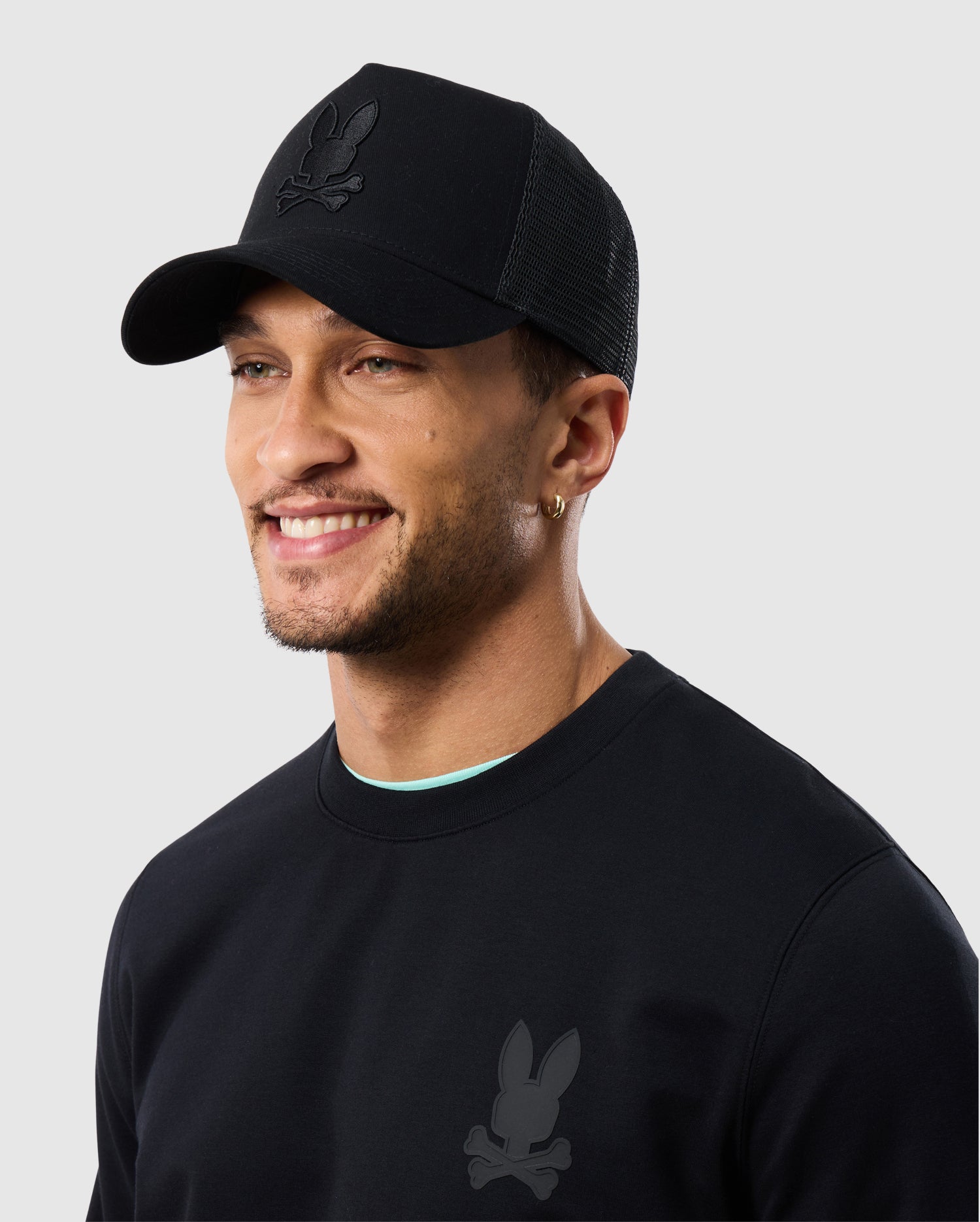 A young man wearing a Psycho Bunny HOUSTON TRUCKER HAT and t-shirt with a small rabbit logo, smiling slightly, against a light grey background.