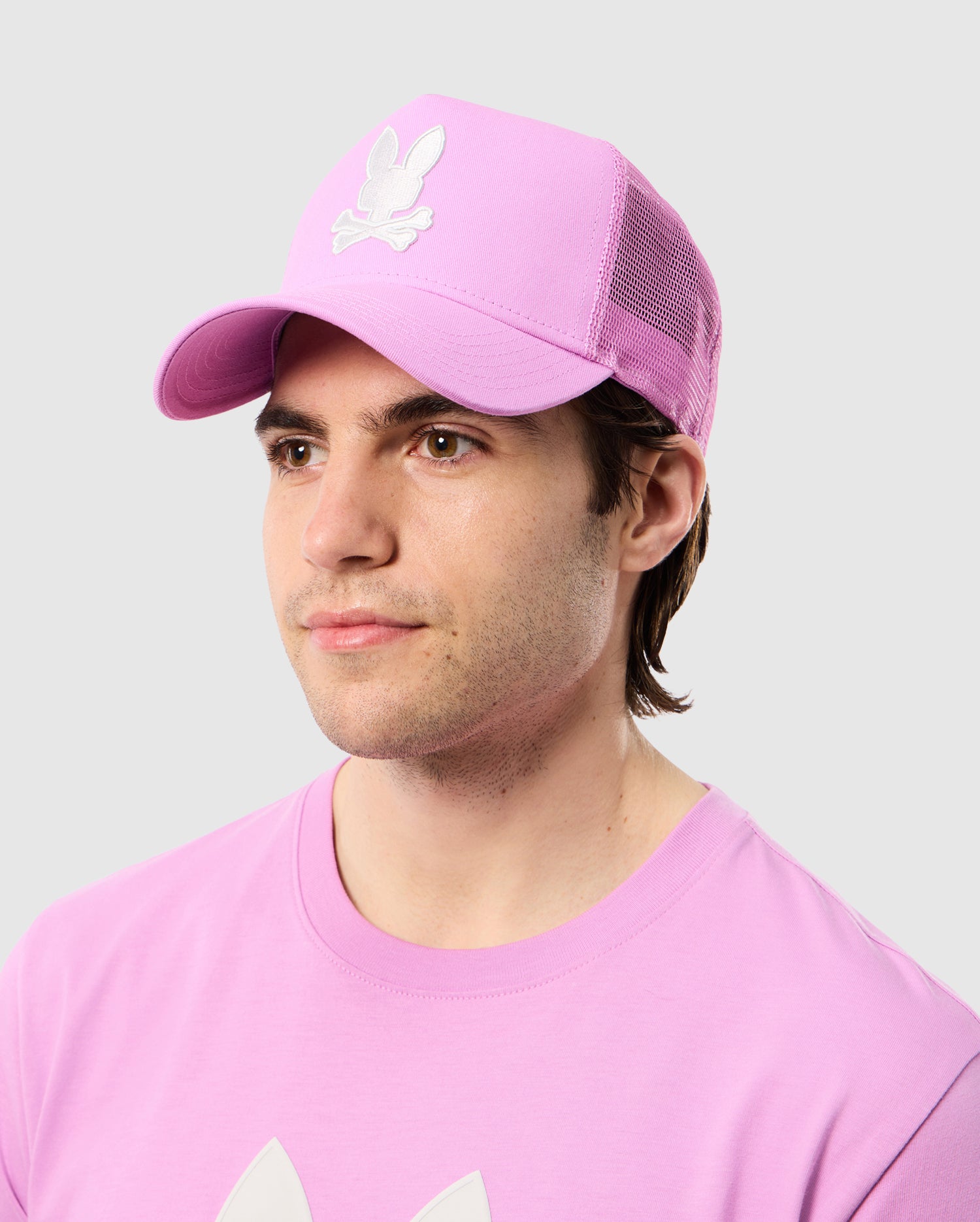 A man wearing a pink t-shirt and a matching pink Psycho Bunny Houston trucker cap with embroidered eyelet vents and a white rabbit logo on it, looking to his left against a light gray background.