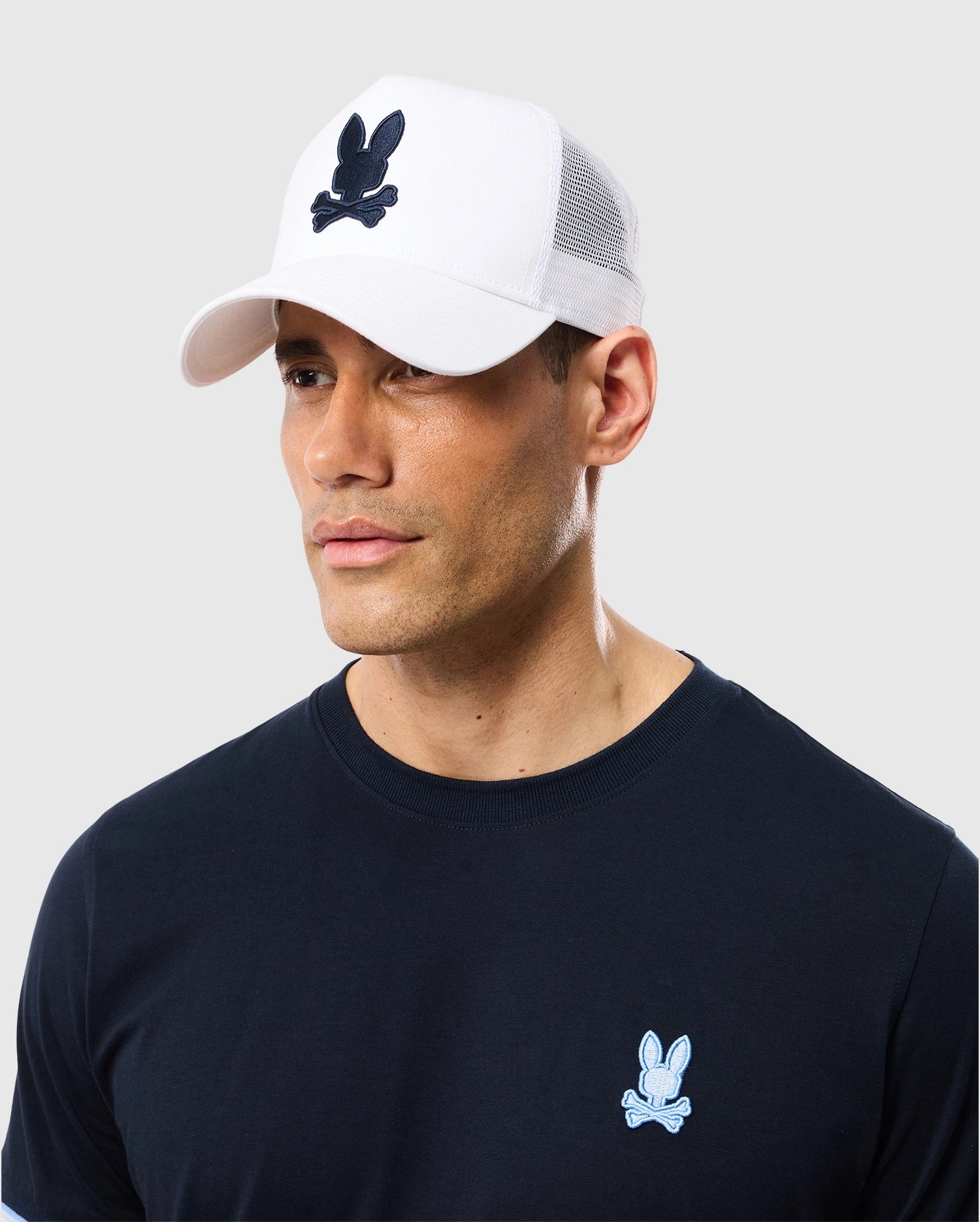 A man wearing a white and black Psycho Bunny Houston trucker cap with a rabbit emblem and embroidered eyelet vents, alongside a dark blue t-shirt adorned with a matching rabbit logo on the chest.