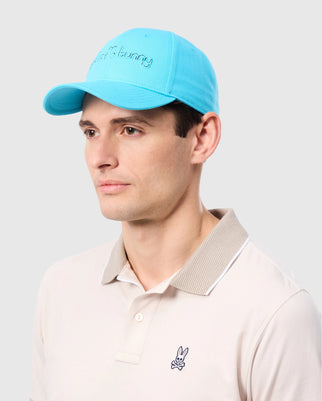 A person is wearing the Men's Louise Baseball Cap in light blue cotton twill from Psycho Bunny and a beige polo shirt with a small embroidered bunny logo on the chest. The cap features an intricate embroidered design. The person is gazing to the right against a plain gray background.