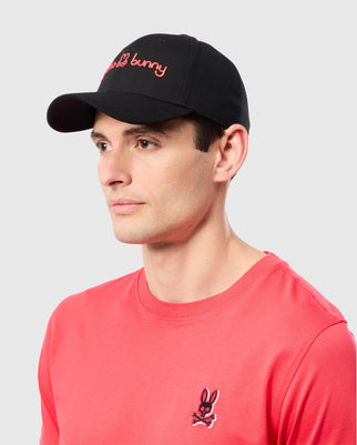 A person wearing the MENS LOUISE BASEBALL CAP - B6A551C200, a black cotton twill baseball cap from Psycho Bunny with red embroidered text and a black bunny logo, paired with a red t-shirt that features the same black bunny logo on the chest. The background is plain white.