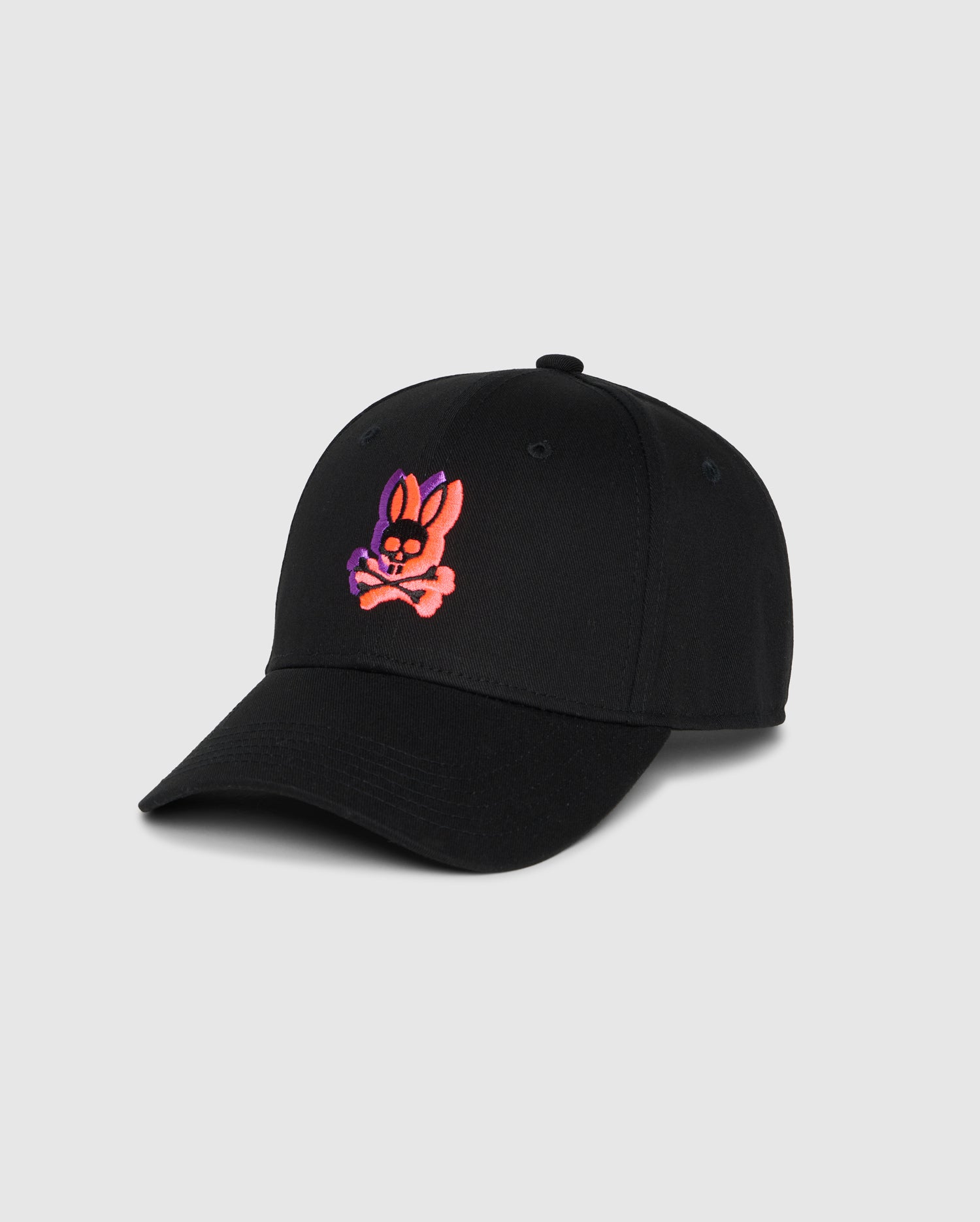 A black Psycho Bunny MENS GROVES BASEBALL HAT - B6A556C2HT featuring a small embroidered design of a cartoonish pink and purple bunny face over crossed bones on the front. The snapback baseball hat is displayed against a plain light grey background.