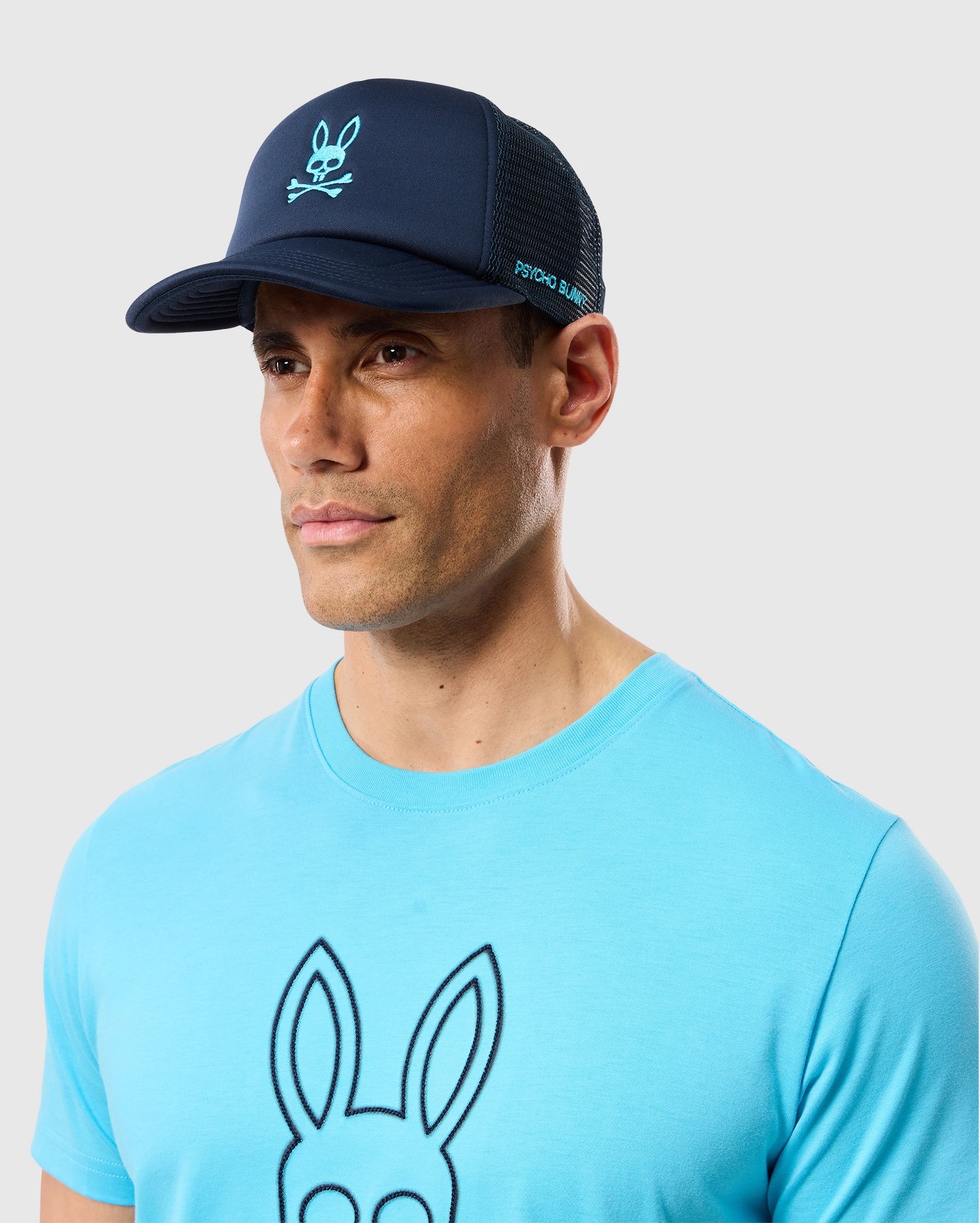 A man wearing a blue Psycho Bunny t-shirt and a navy blue Psycho Bunny MENS PARIS TRUCKER CAP - B6A591C200 with bunny logo mesh paneling. The background is plain light gray.