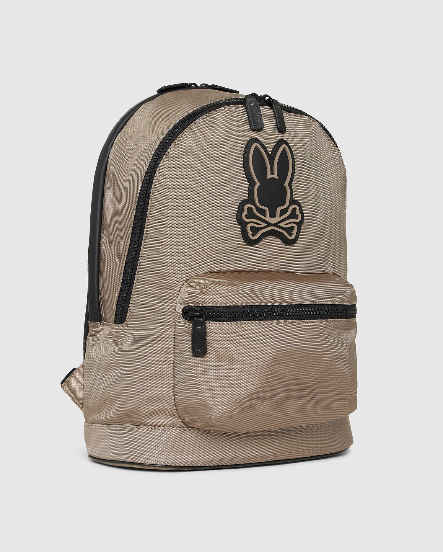 A tan MENS DOME BACKPACK - B6A628Z1BP by Psycho Bunny features a black bunny logo with crossbones on the front. It boasts a large main compartment, a padded laptop compartment, and a smaller front zippered pocket, all accented with black zippers and straps. The background is plain white.