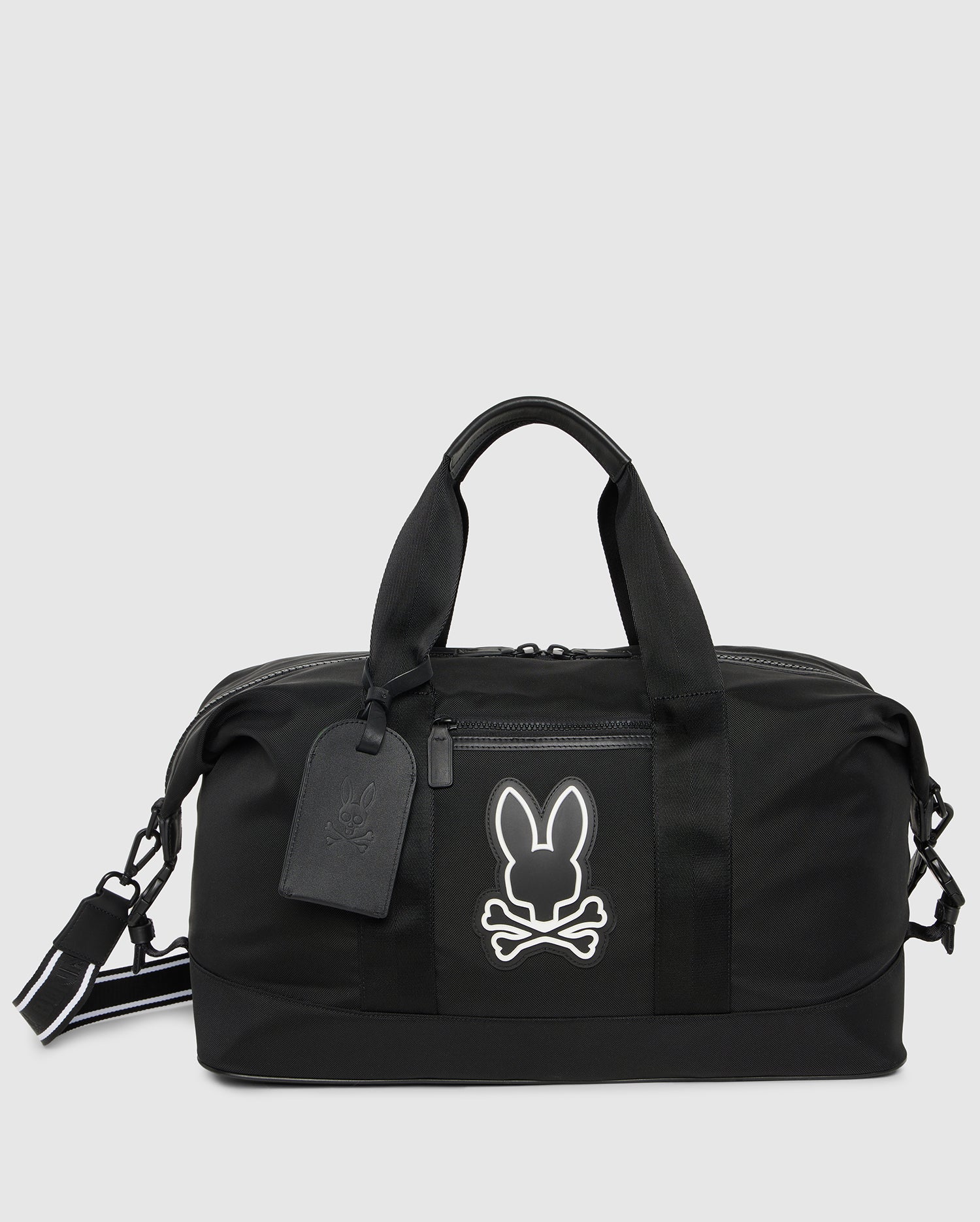 A black MENS WEEKENDER BAG - B6A629Z1BG by Psycho Bunny featuring a white outline of a bunny skull and crossbones design on the front. Perfect as travel luggage, the bag has two handles, a detachable shoulder strap, a zipper closure, and a small attached luggage tag.