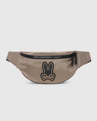 A beige MENS BELT BAG - B6A631Z1BP from Psycho Bunny with a black zipper situated at the top. On the front, there is a rubber bunny patch featuring a stylized bunny head above crossbones, adding a playful yet edgy detail to the design. The pack has an adjustable strap for a comfortable fit.