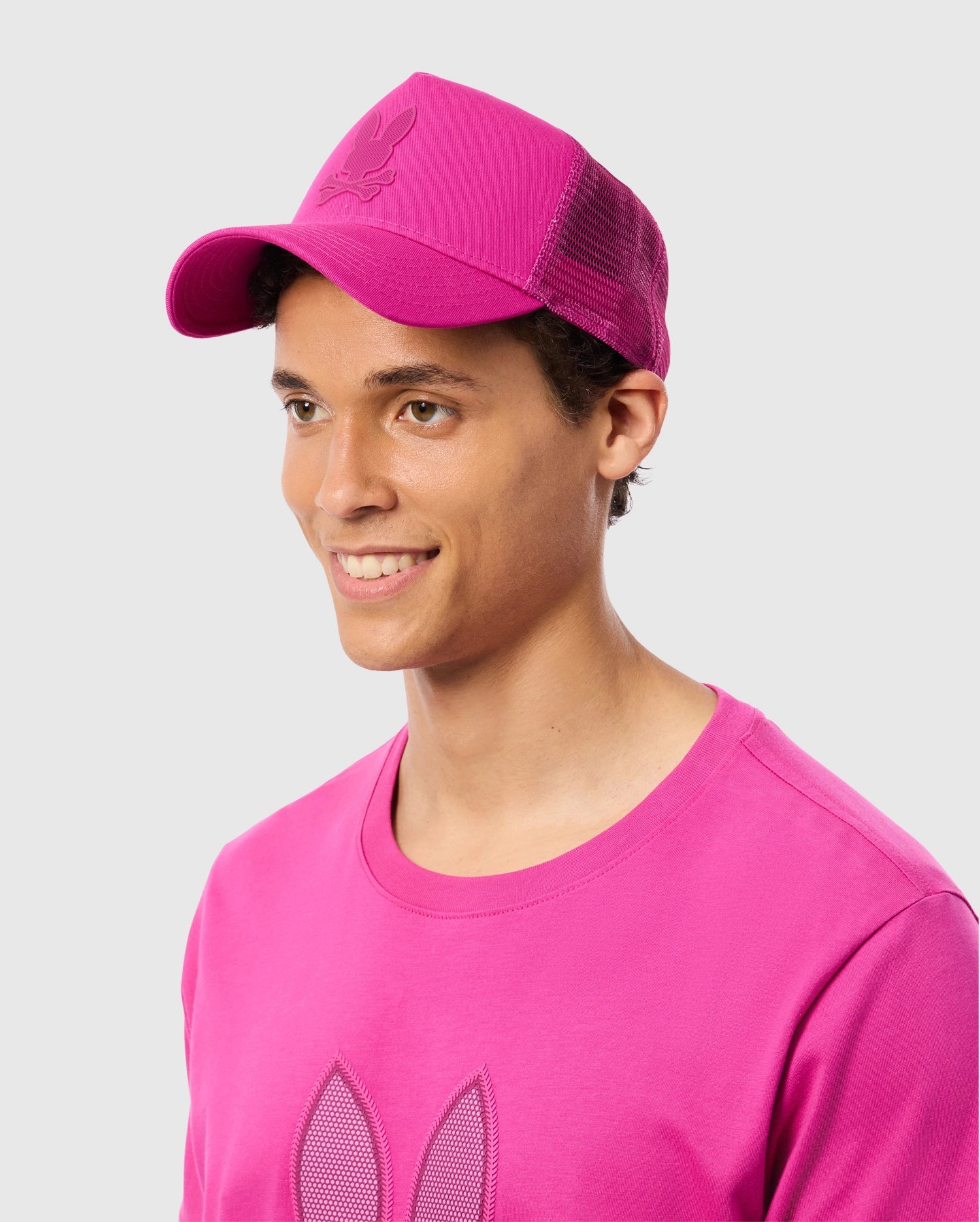 A man in a pink shirt and a Psycho Bunny MENS RIVIERA TRUCKER CAP - B6A667C200 with snapback fastening.