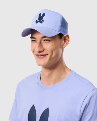 A person smiles while wearing a light purple T-shirt and a matching Psycho Bunny MENS RIVIERA TRUCKER CAP - B6A667C200. Both the cap and shirt feature a playful black bunny emblem, evoking early 2000s nostalgia. The background is plain light gray.