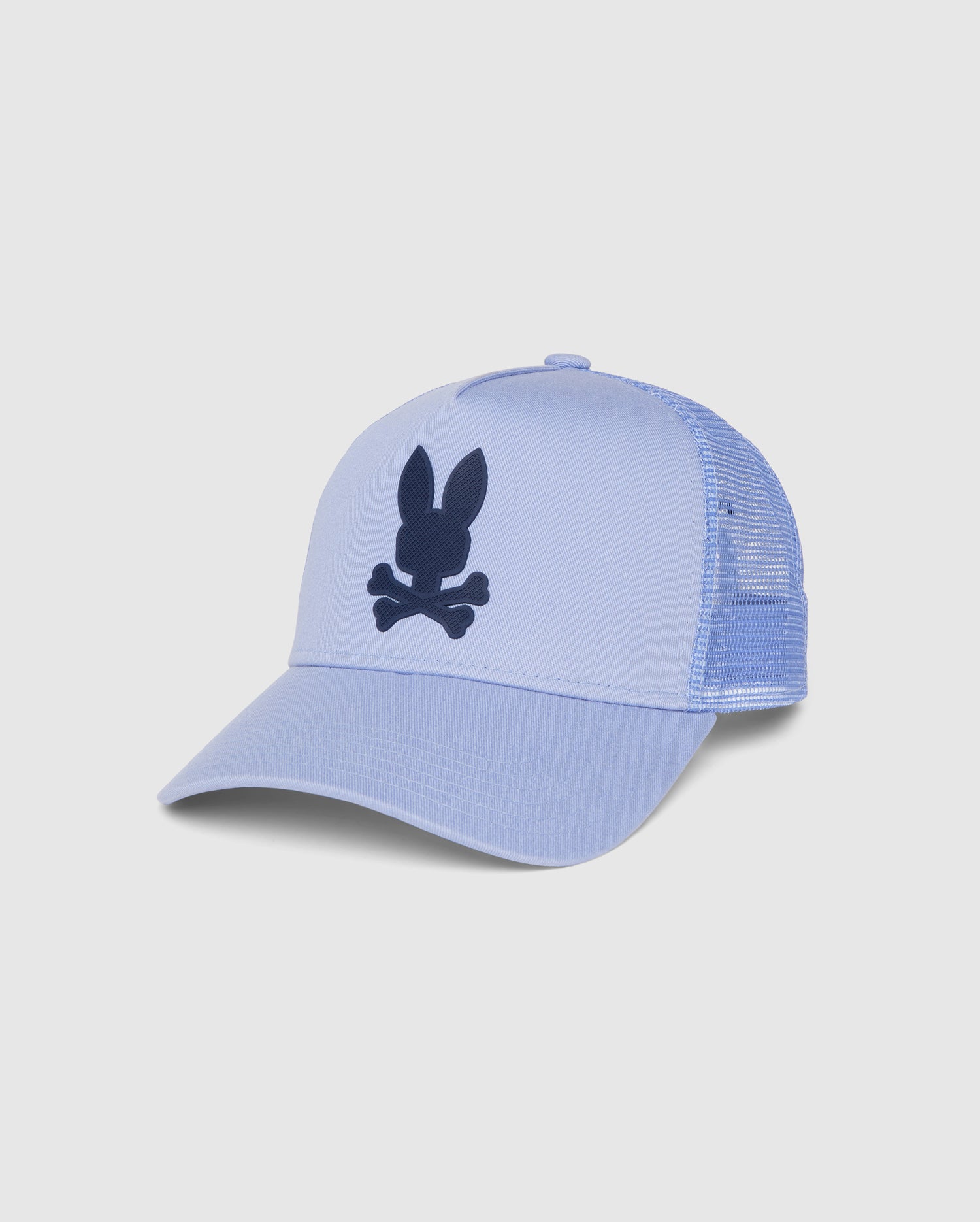 A light blue MENS RIVIERA TRUCKER CAP - B6A667C200 with a mesh back and a solid front panel. The front features a black graphic of a bunny head with crossbones beneath it, capturing early 2000s nostalgia. The cap has a curved brim and an adjustable snapback fastening at the back by Psycho Bunny.