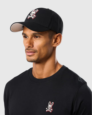 A man wearing the Psycho Bunny MENS KAYDEN BASEBALL CAP - B6A677C200, which features cotton twill material, a white underside, and an embroidered Bunny logo with a skull and crossbones. He has a goatee and is also wearing a black long-sleeved shirt adorned with the same Bunny logo on the chest. The background is light gray.