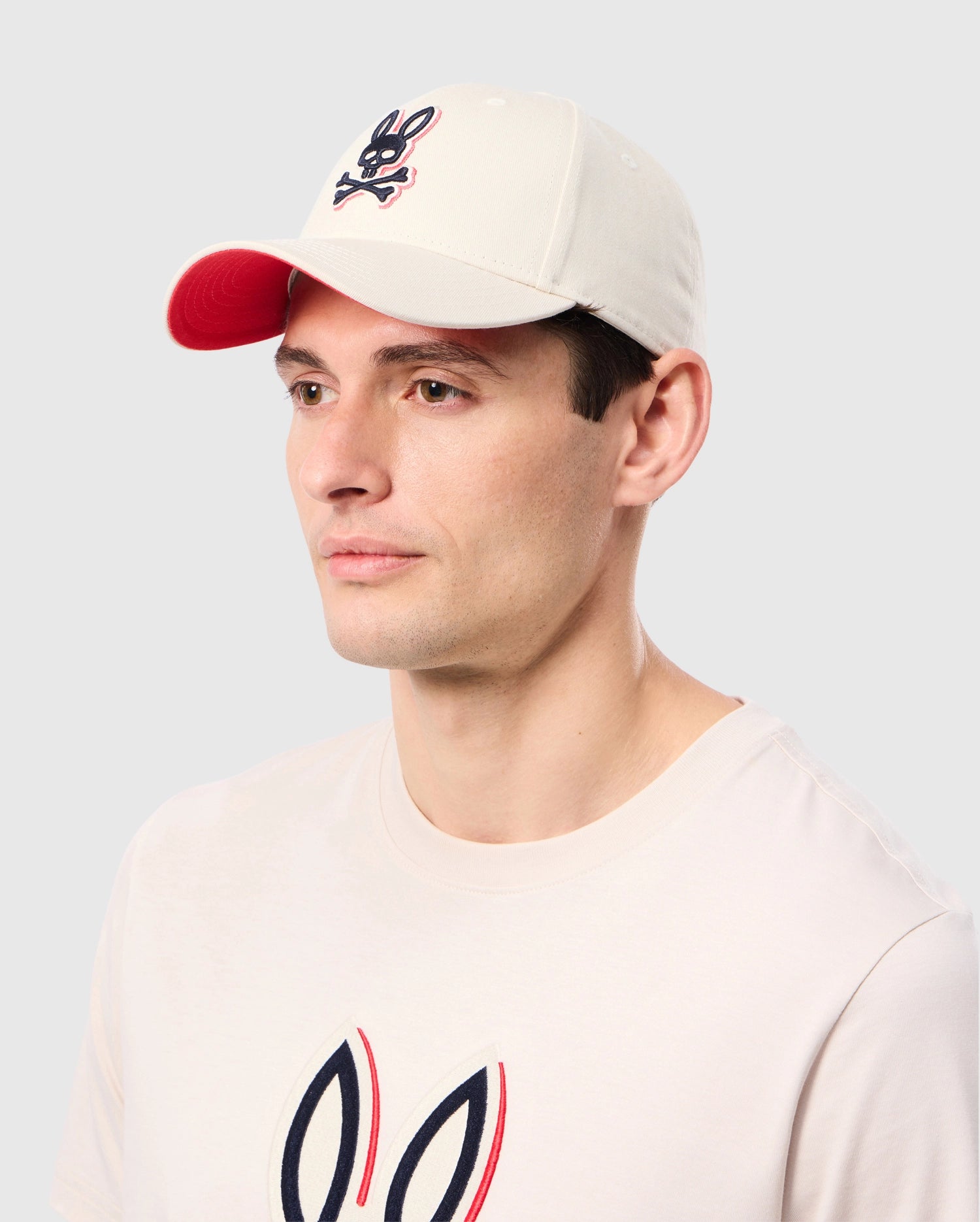 A man is wearing a white MENS KAYDEN BASEBALL CAP - B6A677C200 from Psycho Bunny, featuring an embroidered Bunny logo with a black and white rabbit skull on the front. He is dressed in a matching white T-shirt with the same design. The durable cotton twill cap stands out against the plain, light gray background.