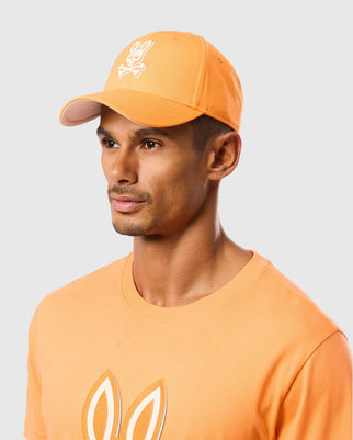 A person is shown wearing a Psycho Bunny MENS KAYDEN BASEBALL CAP - B6A677C200 in orange cotton twill, featuring a white bunny logo with a skull and bunny ears on the front. They are also dressed in an orange T-shirt with a matching bunny ear graphic. The background is plain light gray.