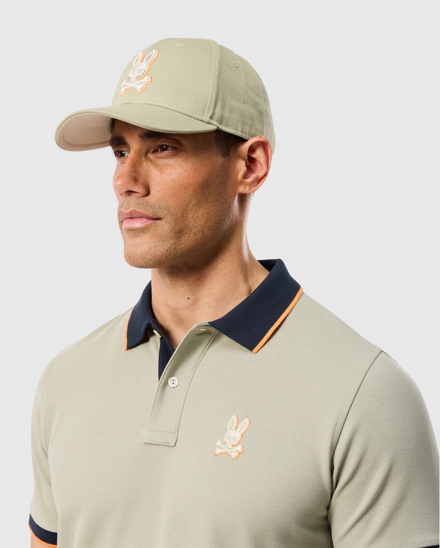 A man wearing a light green Psycho Bunny MENS KAYDEN BASEBALL CAP - B6A677C200 and a matching polo shirt with a bunny logo on both. He is facing slightly to the right, and the background is plain white. The shirt has a dark collar with an orange trim.