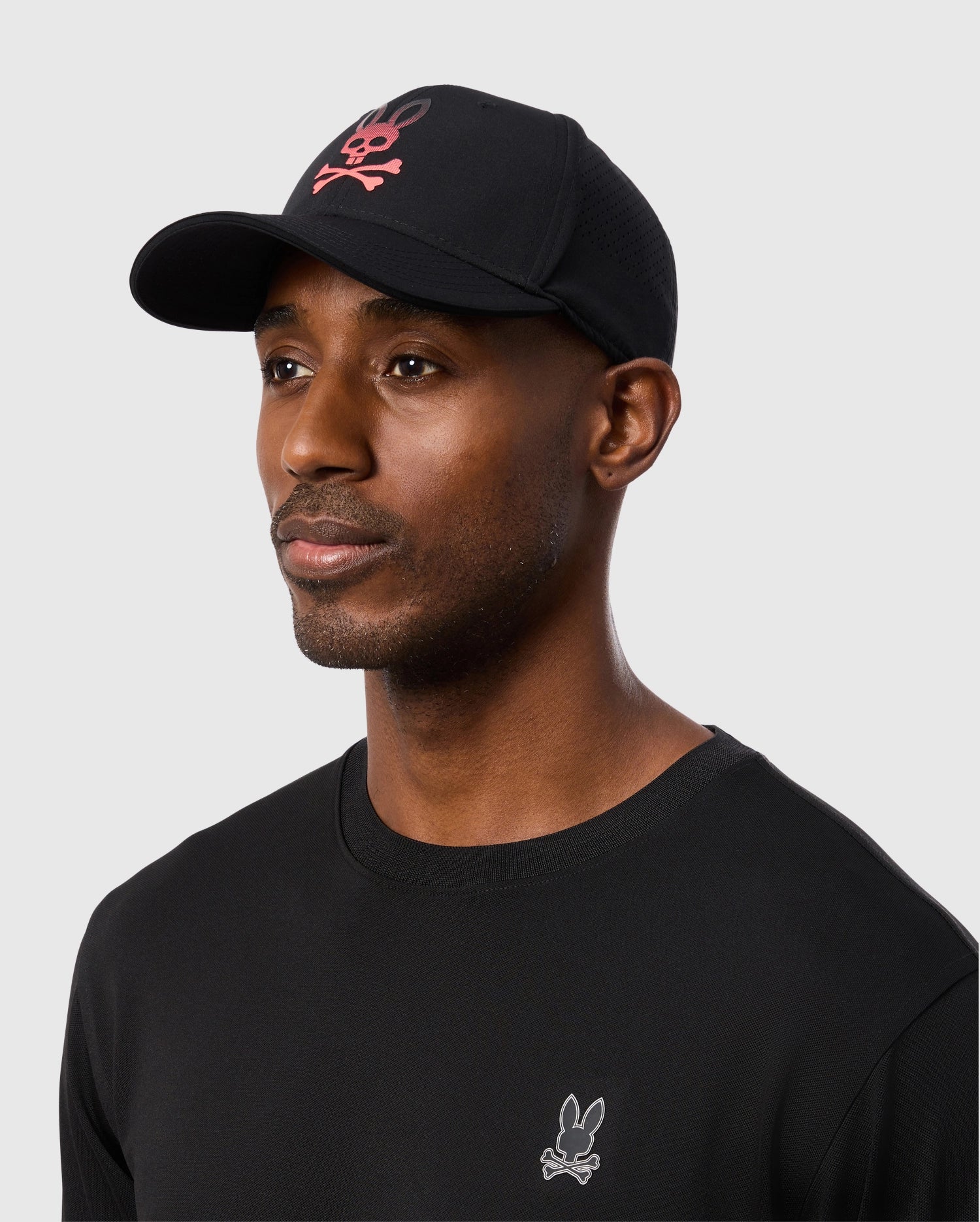 Maxwell wore a 100% cotton black long-sleeve shirt with a small white bunny logo on the chest and topped it off with the MENS MAXWELL SPORT CAP - B6A783D200 by Psycho Bunny, featuring a red skull and crossbones emblem. The background was plain gray.