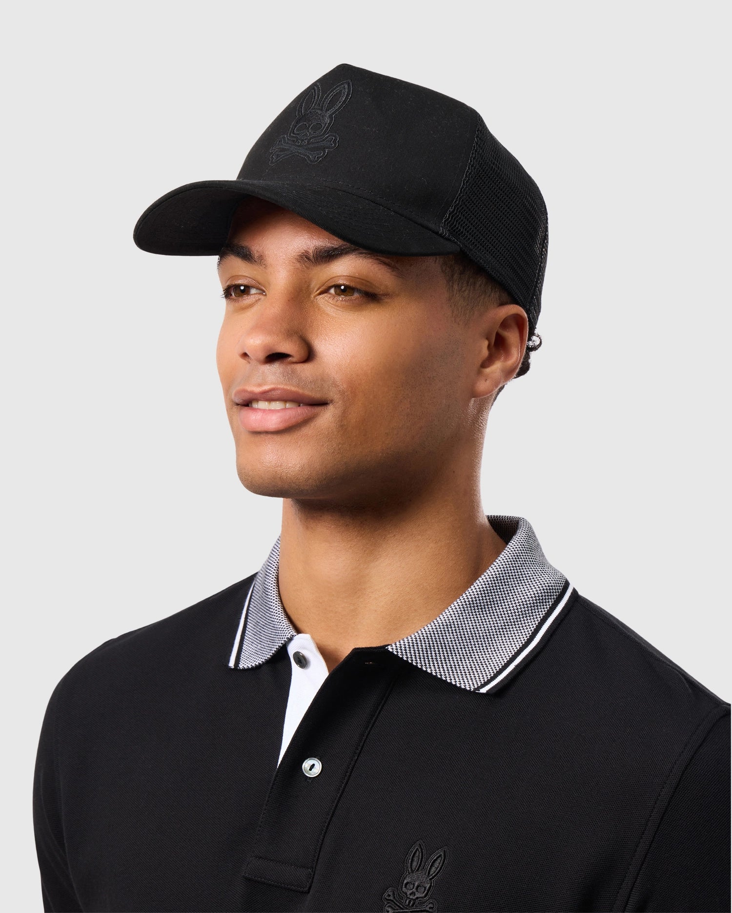 A man is wearing a Psycho Bunny MENS MALTON TRUCKER CAP - B6A797D200 featuring an embroidered design on the front panel and mesh ventilation at the back. He is also dressed in a black collared shirt with white and gray trim. The background is plain and light gray.