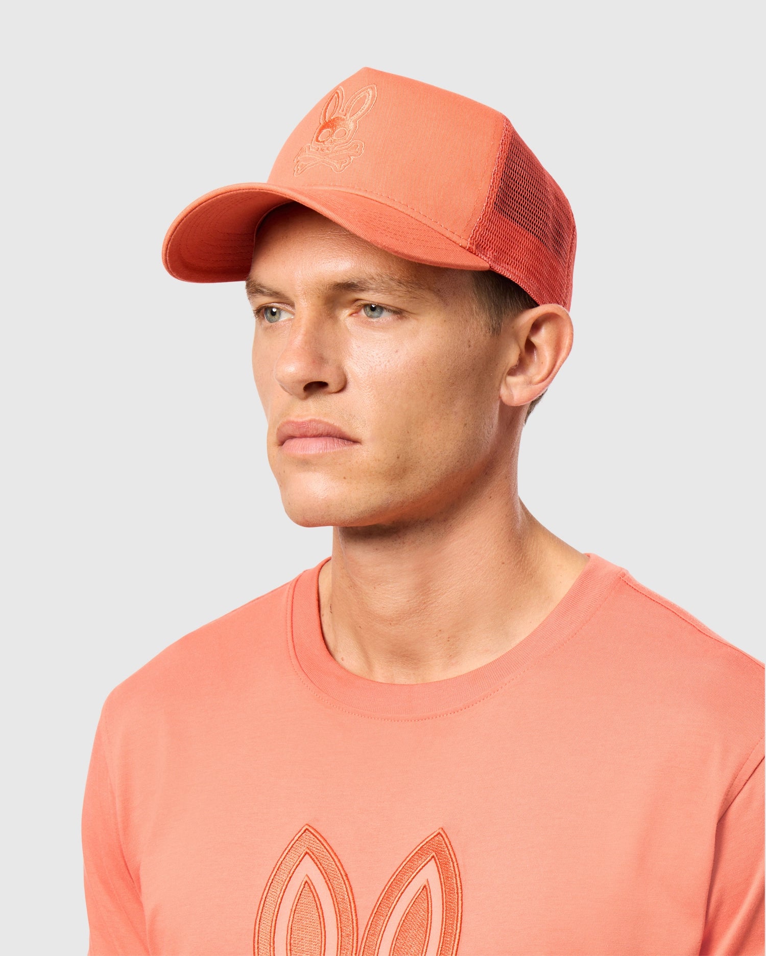 A person is wearing a MENS MALTON TRUCKER CAP from Psycho Bunny, featuring coral mesh ventilation and an embroidered logo on the front, accompanied by a matching t-shirt with a similar chest design. Set against a plain background, their neutral expression conveys an effortlessly active lifestyle.