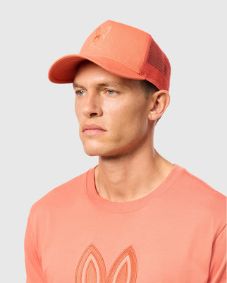 A person is wearing a MENS MALTON TRUCKER CAP from Psycho Bunny, featuring coral mesh ventilation and an embroidered logo on the front, accompanied by a matching t-shirt with a similar chest design. Set against a plain background, their neutral expression conveys an effortlessly active lifestyle.