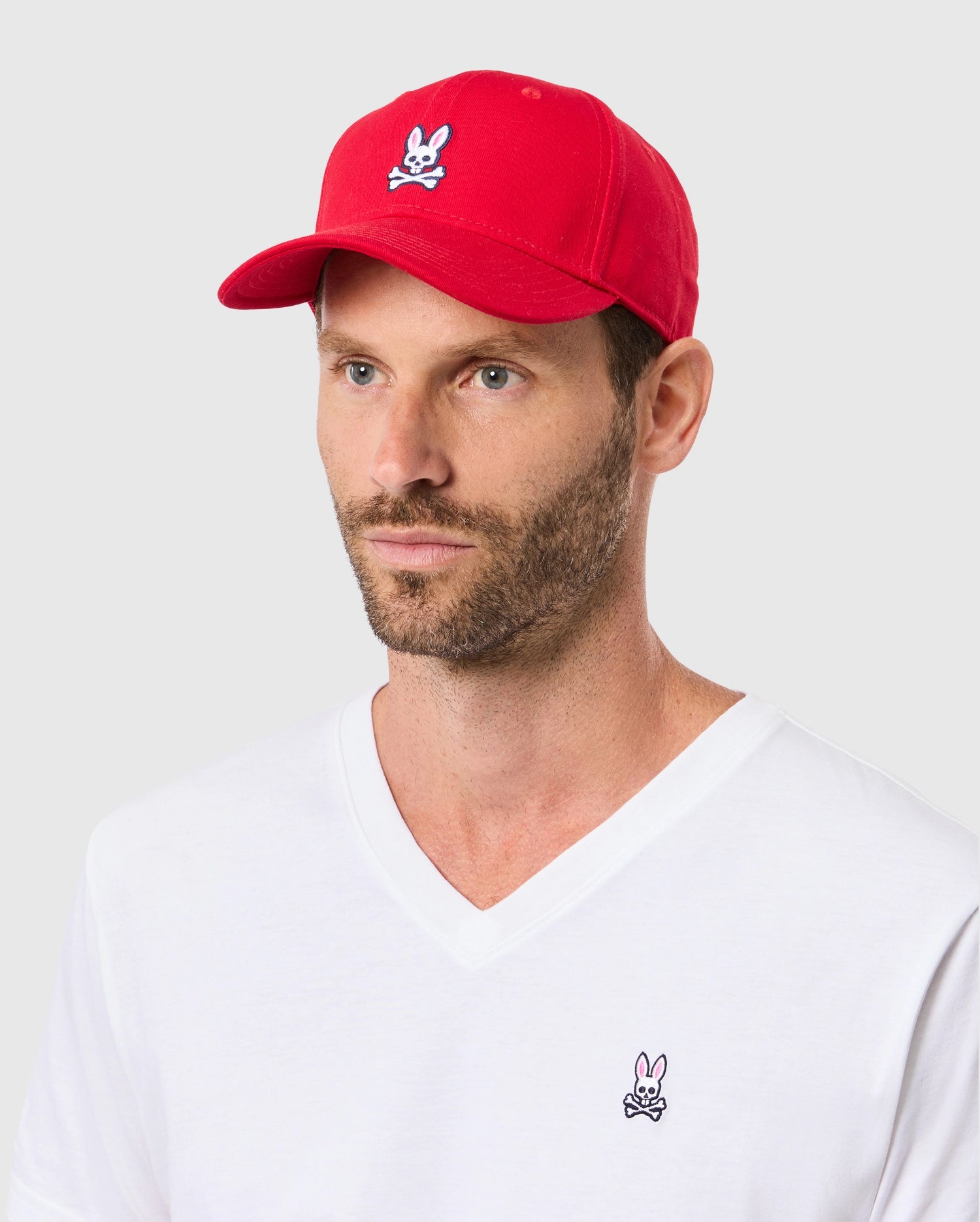 A person wearing the Psycho Bunny MENS CLASSIC BASEBALL CAP (B6A816A1HT) in red cotton twill with an adjustable fit, featuring a white rabbit skull logo, perfectly matches their white V-neck T-shirt with the same design against a simple light gray backdrop.