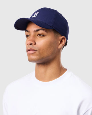 A person wearing a Psycho Bunny MENS CLASSIC BASEBALL CAP - B6A816A1HT in timeless navy blue, featuring a white bunny logo on the front, looking slightly to the side. The individual is also dressed in a plain white 100% cotton crew-neck shirt against a light grey background.