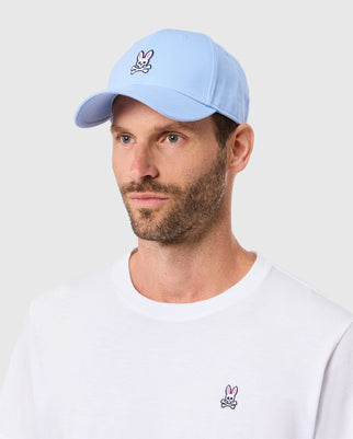 A man dons the Psycho Bunny MENS CLASSIC BASEBALL CAP - B6A816B200 in a light blue hue, crafted entirely from cotton and embellished with the distinctive bunny logo. It complements his white T-shirt featuring the same emblem against a neutral gray background, highlighting the ensemble's understated elegance.