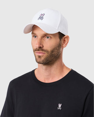 A bearded man wears a Psycho Bunny classic white cotton baseball cap (product code: B6A816A1HT) and a sleek black T-shirt, both adorned with a small embroidered logo of a rabbit above crossed bones. He looks off to the side against a simple background.