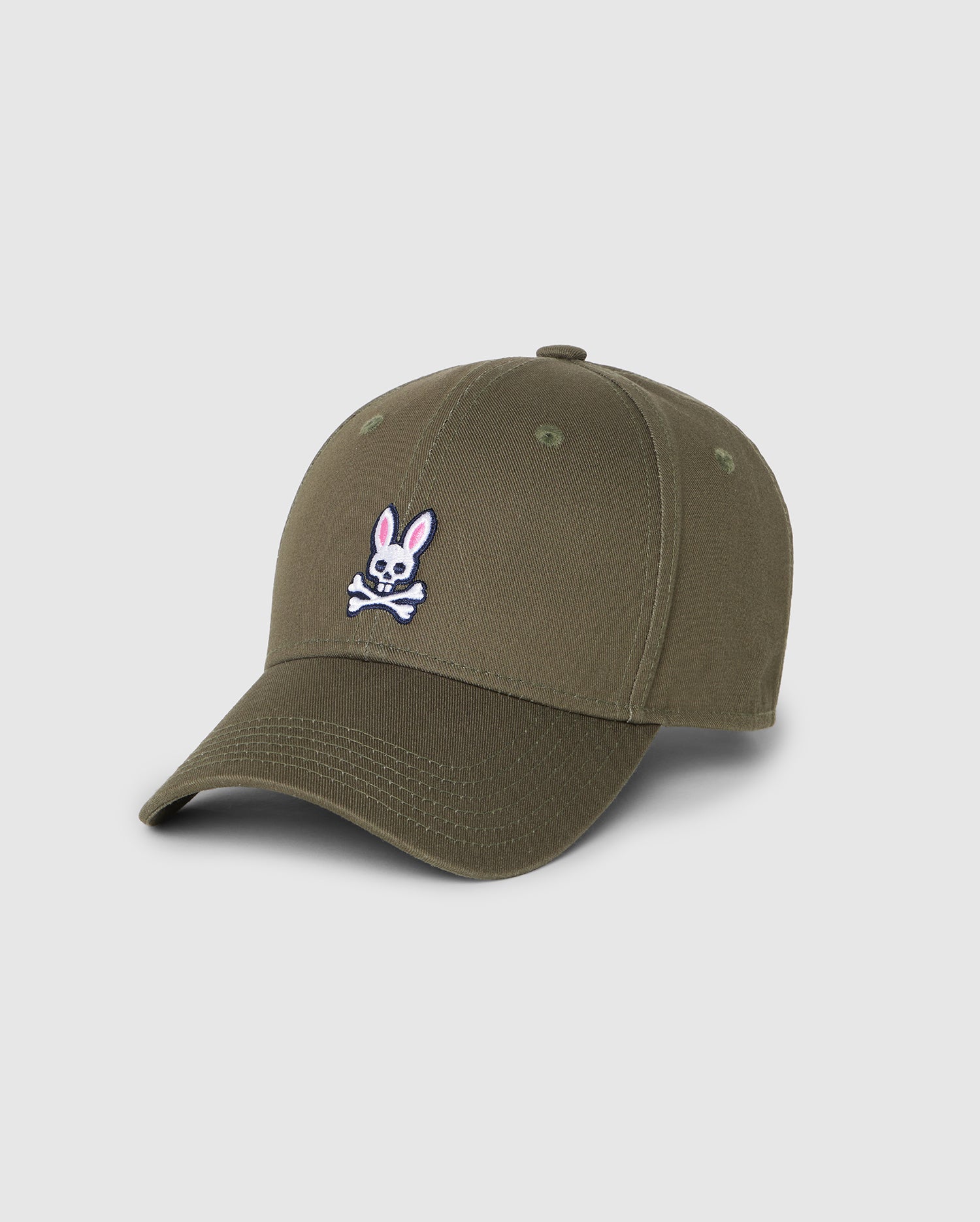 A men's classic baseball cap, the MENS CLASSIC BASEBALL CAP - B6A816B200 by Psycho Bunny, is made from 100% cotton and features an embroidered design of a white and pink cartoon rabbit above crossed bones on the front. The cap has a curved brim, ventilation holes on the top, and a plain white background.