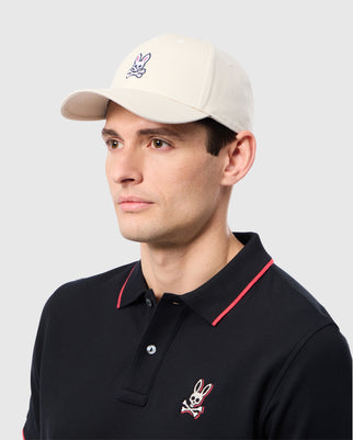 A person wearing the MENS CLASSIC BASEBALL CAP - B6A816B200 by Psycho Bunny, a cream-colored hat featuring a bunny logo and crafted from 100% cotton, poses against a light gray background while pairing it with a black polo shirt accented in red for a casual yet stylish look.