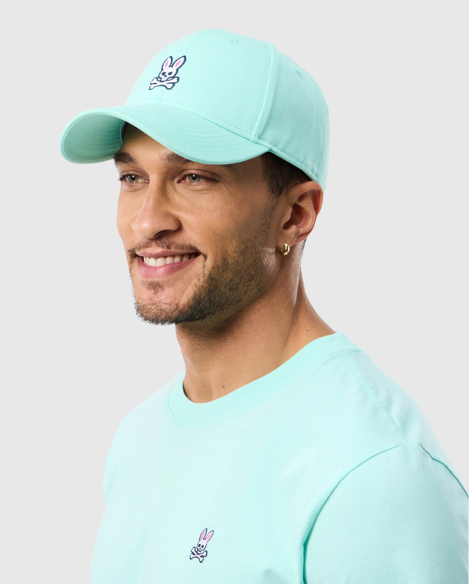 A man wearing a light blue 100% cotton t-shirt with a bunny logo and a MENS CLASSIC BASEBALL CAP - B6A816C200 by Psycho Bunny smiles while looking to the side. He has short hair, a beard, and a small hoop earring in one ear. The cap features snapback adjustability. The background is plain and light-colored.
