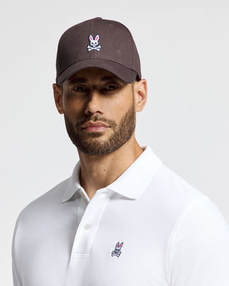 A man with a beard wears the Psycho Bunny Men's Classic Baseball Cap (B6A816D200) in dark cotton twill, adorned with the distinctive white skull and bunny logo. The cap's adjustable fit perfectly complements his white polo shirt, showcasing the same emblem on the chest against a plain light-colored background.