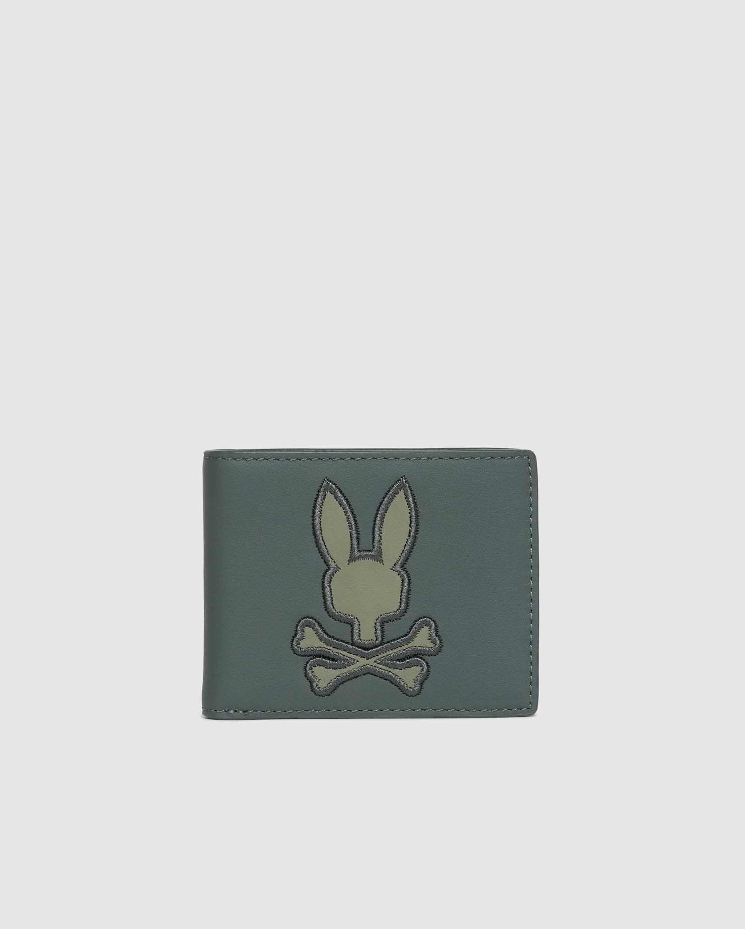 Introducing the MENS WORDMARK LEATHER WALLET - B6A868D200 by Psycho Bunny, a stylish accessory for men. This trendy green leather wallet showcases an embroidered rabbit head above crossbones on the front, set against a sleek white background.