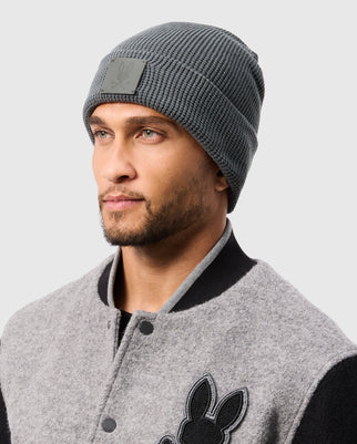 A person wearing the MENS WAFFLE BEANIE in gray from Psycho Bunny and a gray and black jacket featuring the Psycho Bunny logo gazes to the left against a plain background.