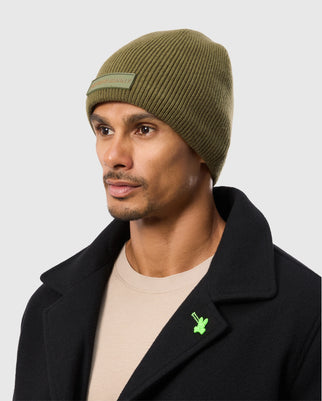 A person is wearing a MENS SPORT BEANIE - B6A874D200 by Psycho Bunny in olive-green, along with a beige shirt and a black coat featuring a green leaf pin. The background is plain gray.