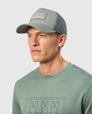 A man wearing a MENS TACOMA TRUCKER CAP - B6A941D200 by Psycho Bunny, featuring bunny silhouettes, pairs it with a complementary t-shirt. He looks to the side against a plain backdrop, effortlessly merging casual style with a playful design.