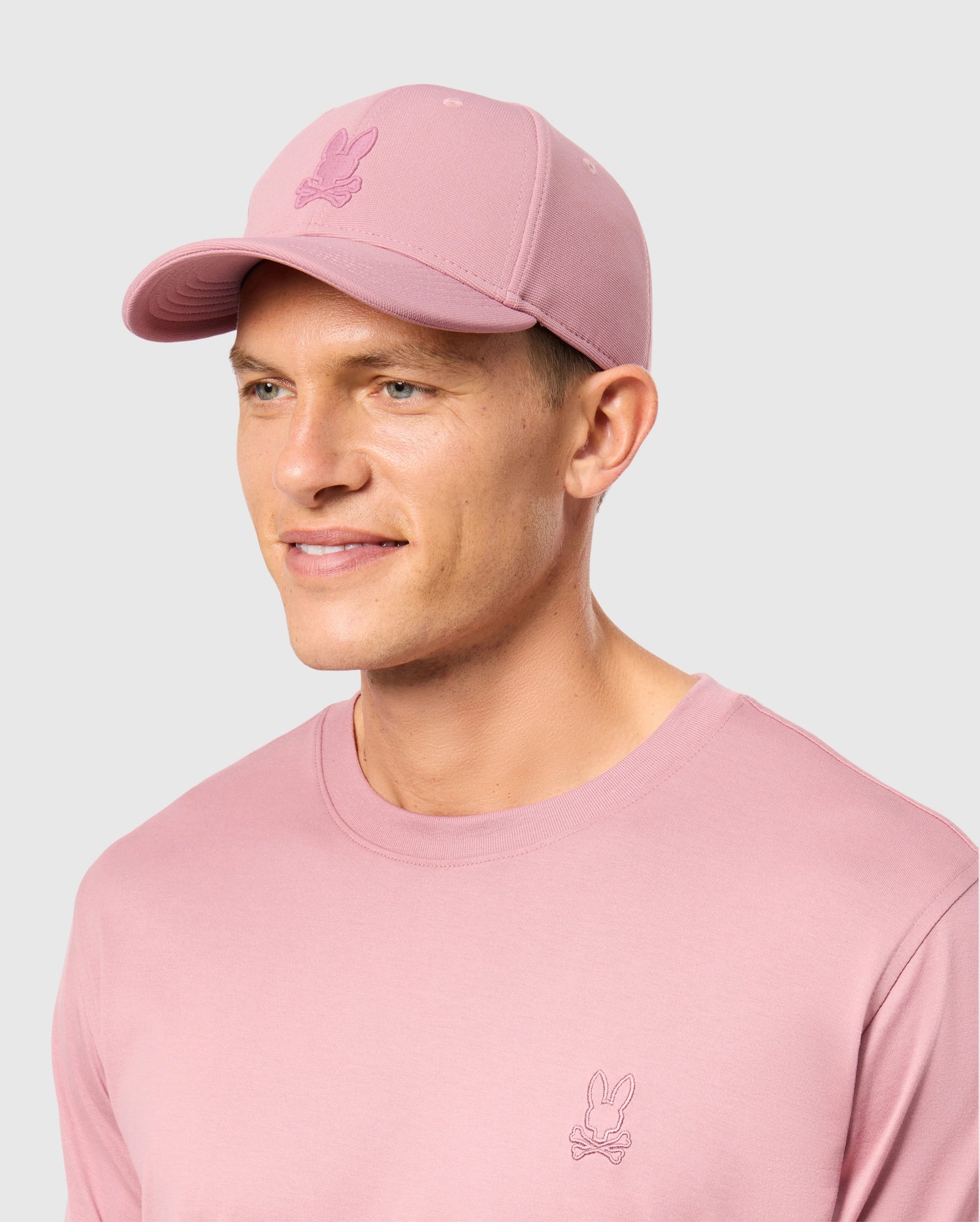 A person wearing a MENS JAMES PREMIUM BASEBALL CAP by Psycho Bunny, along with a matching pink shirt, both showcasing an embroidered Bunny design. The comfortable outfit is crafted from polyester spandex and is set against a plain gray background.