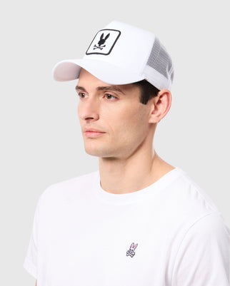 A man is wearing a Psycho Bunny MENS BUNNY PATCH TRUCKER CAP - B6A965C200, featuring a black and white Bunny Patch with a cartoon bunny above crossbones. He is also sporting a white T-shirt with a small pink bunny logo on the chest. The background is plain and light-colored.