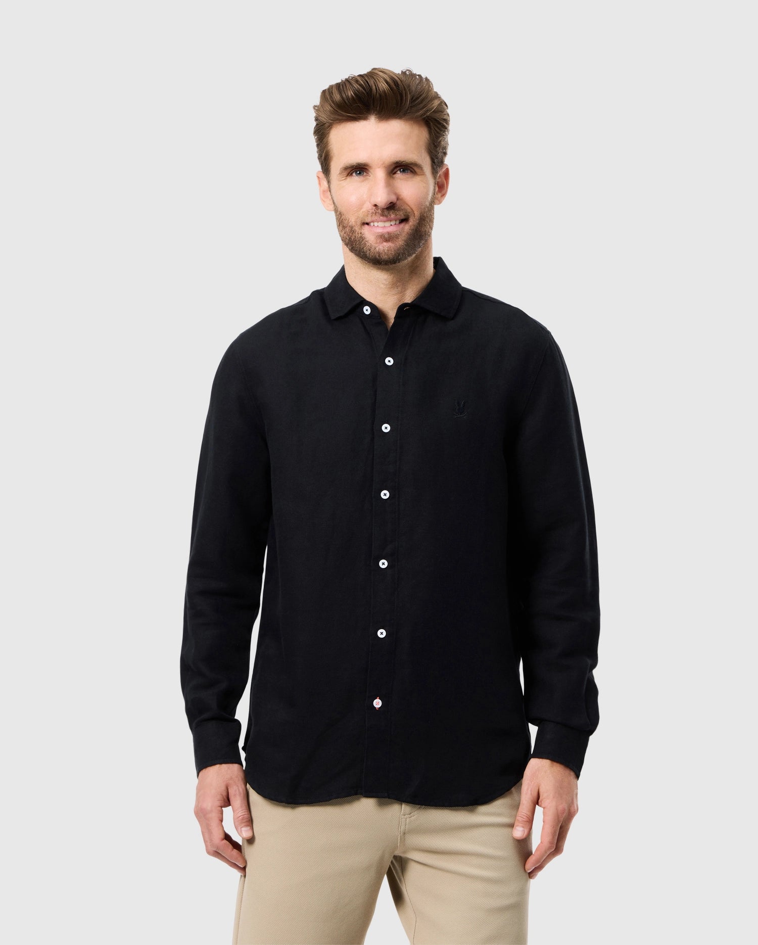 A man with short brown hair and a beard smiles while wearing the Psycho Bunny MENS CAMERON LINEN LONG SLEEVE SHIRT - B6C586C200 with beige pants. He stands against a plain light gray background.