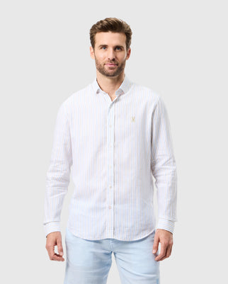 A man with short hair and a trimmed beard is wearing a long-sleeved, light-colored striped linen shirt and light blue pants. He stands against a plain, light gray background, looking directly at the camera with a subtle smile. The MENS WEST LINEN LONG SLEEVE SHIRT - B6C587C200 by Psycho Bunny has a small embroidered emblem.